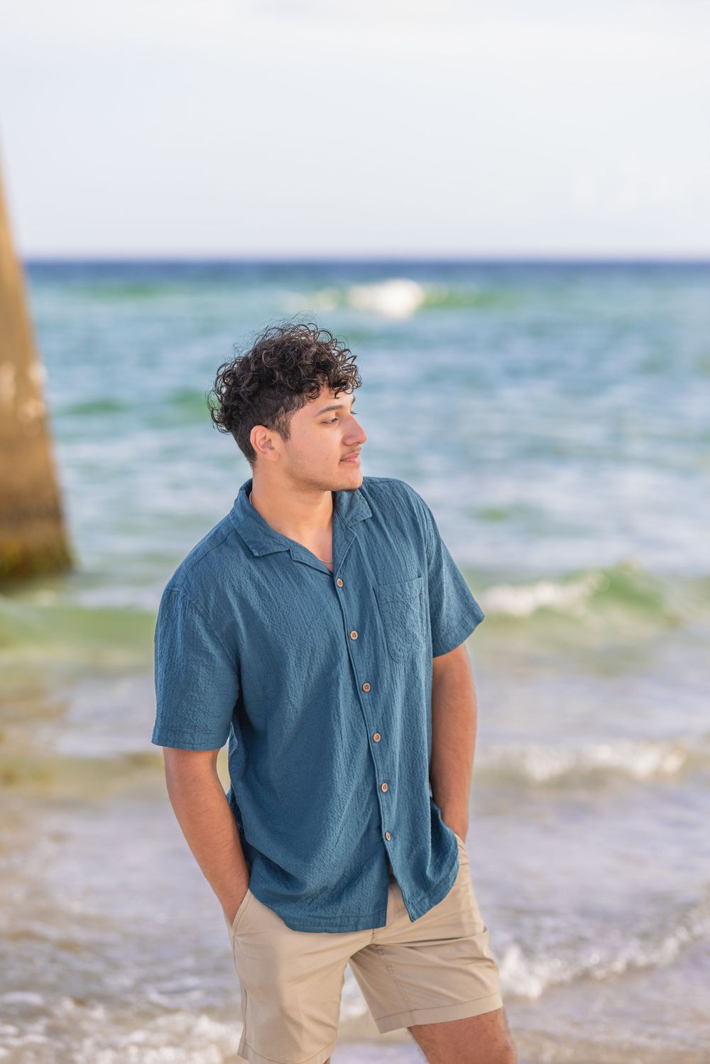 A person in a blue shirt and khaki shorts stands against an ocean backdrop with waves and sunlit water.