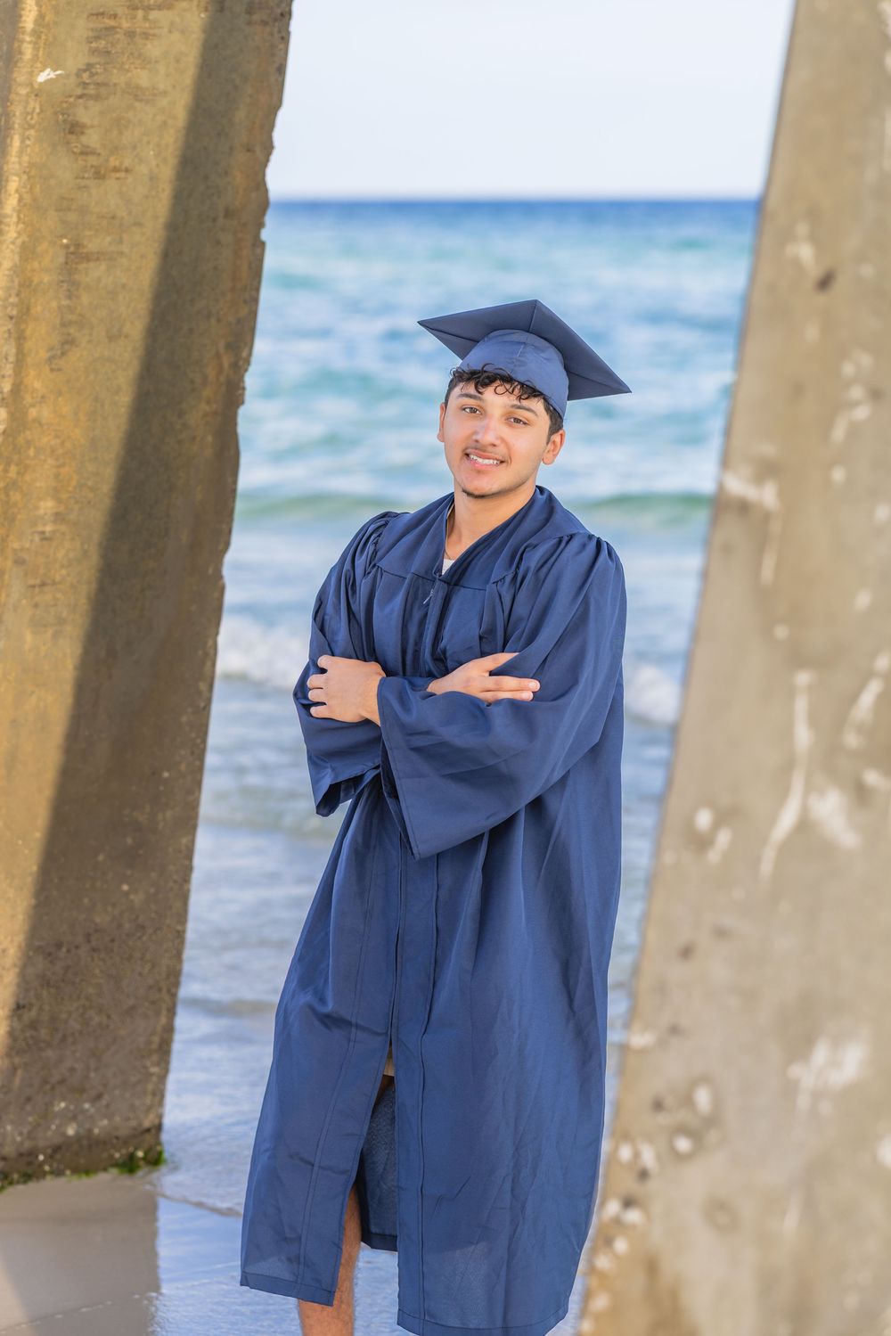 A graduate in navy blue cap and gown poses confidently between concrete pillars on a sunny beach.