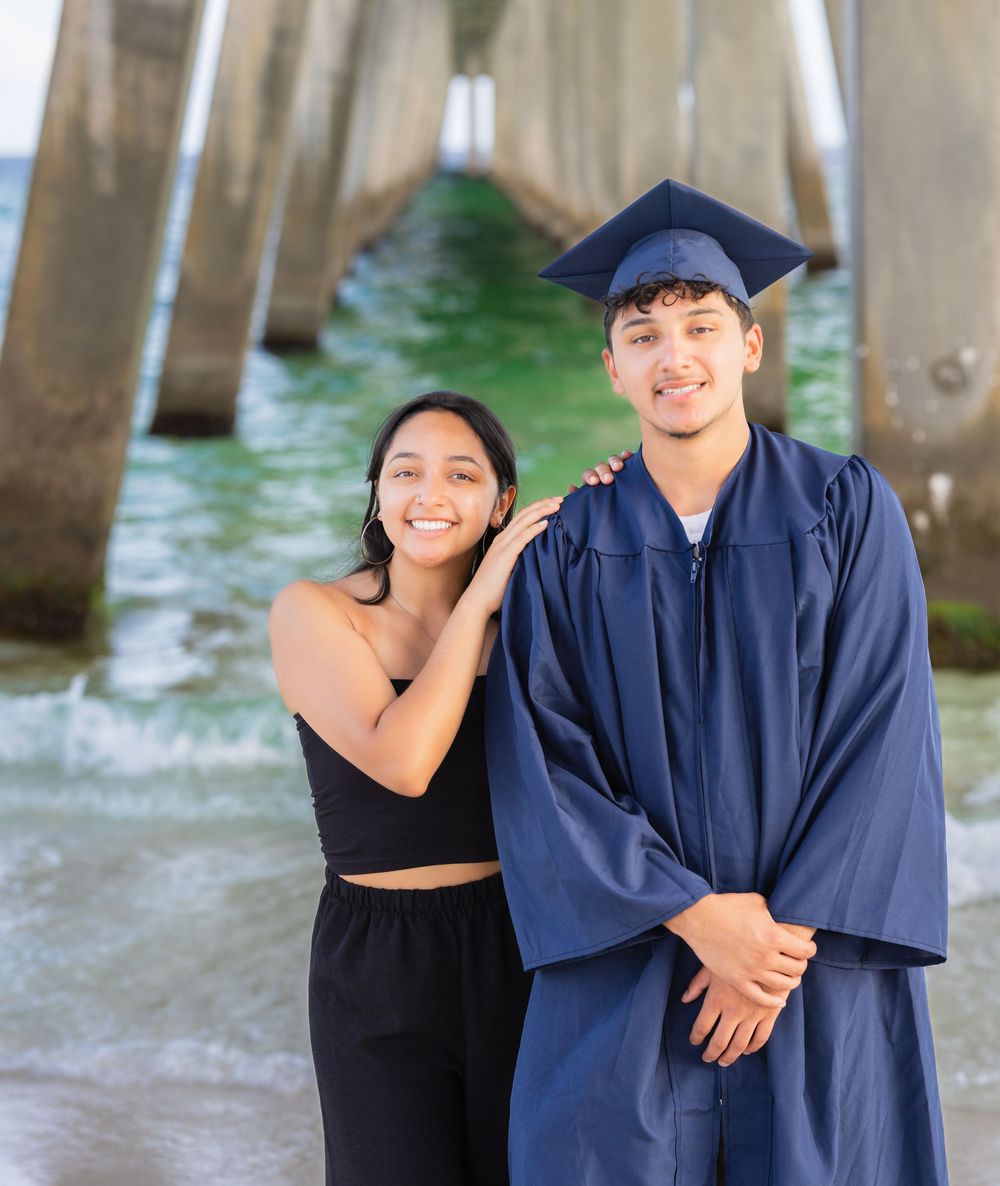 Graduation photos showing a couple by wooden pier posts, with one wearing a navy cap and gown and other in black attire.