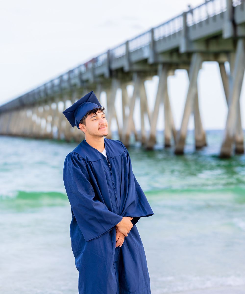 Graduate in navy blue cap and gown stands thoughtfully by a fishing pier on a bright sunny day at the beach.