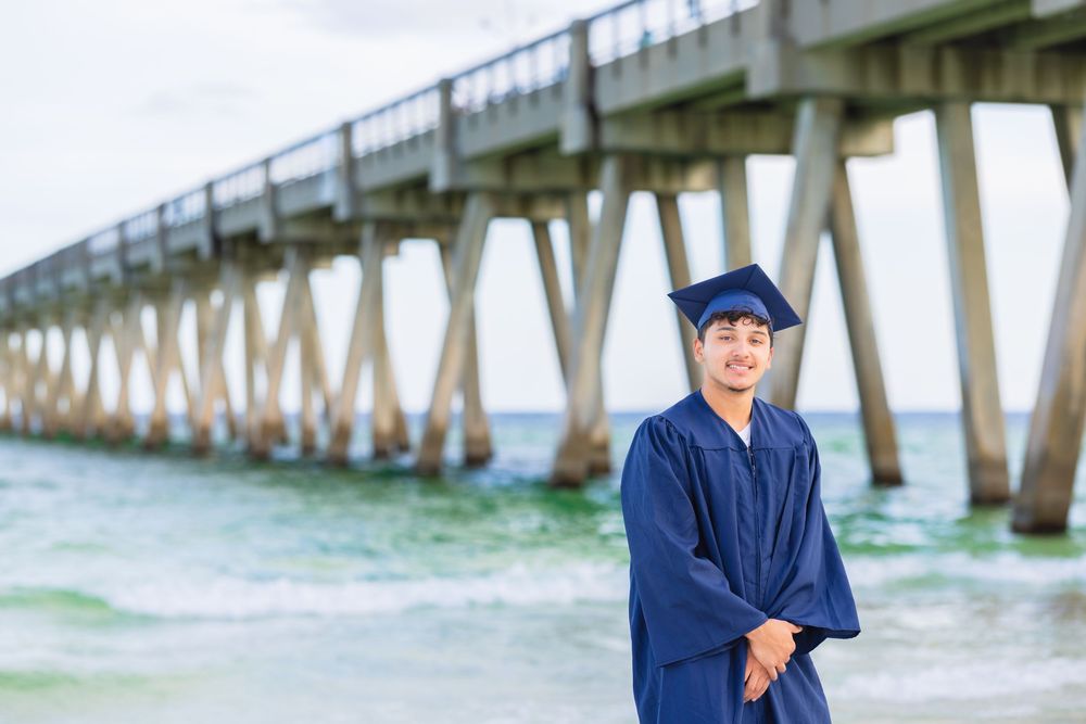 Graduate in blue cap and gown poses near a pier with ocean waves and wooden supports in the background.