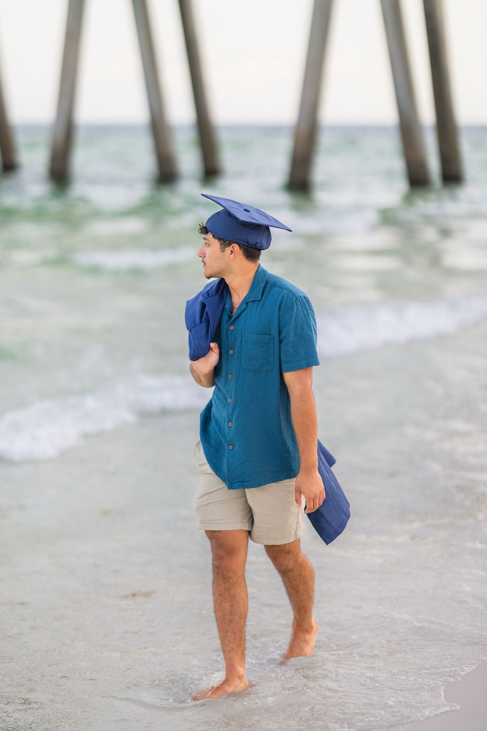 Recent graduate in blue cap and gown walks barefoot on beach with pier in background at sunset.