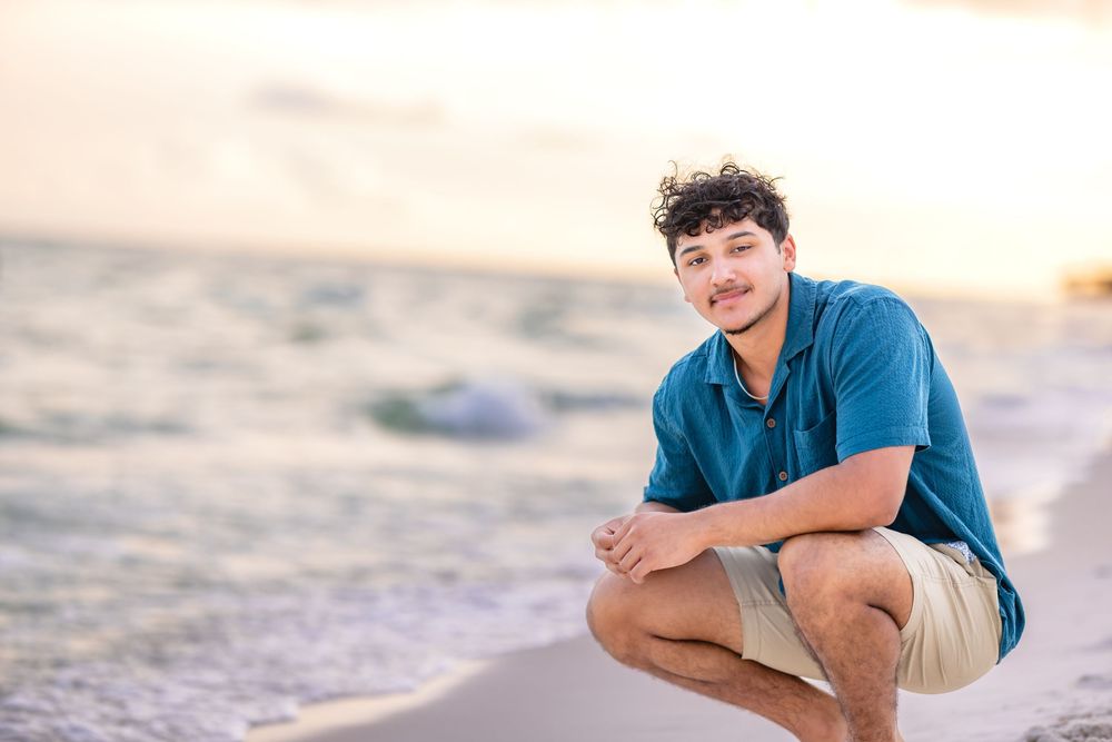 Someone in a blue shirt and khaki shorts crouches on a sandy beach at sunset with ocean waves in the background.