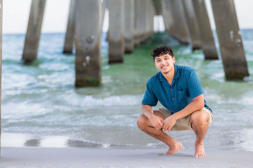 Person in blue shirt crouches on beach near pier pilings with ocean waves and teal water in background.