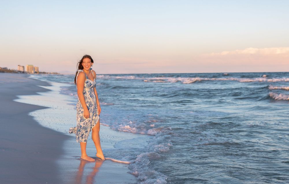 A person in a floral dress walks barefoot along a sandy beach at sunset with ocean waves and shore in the background.