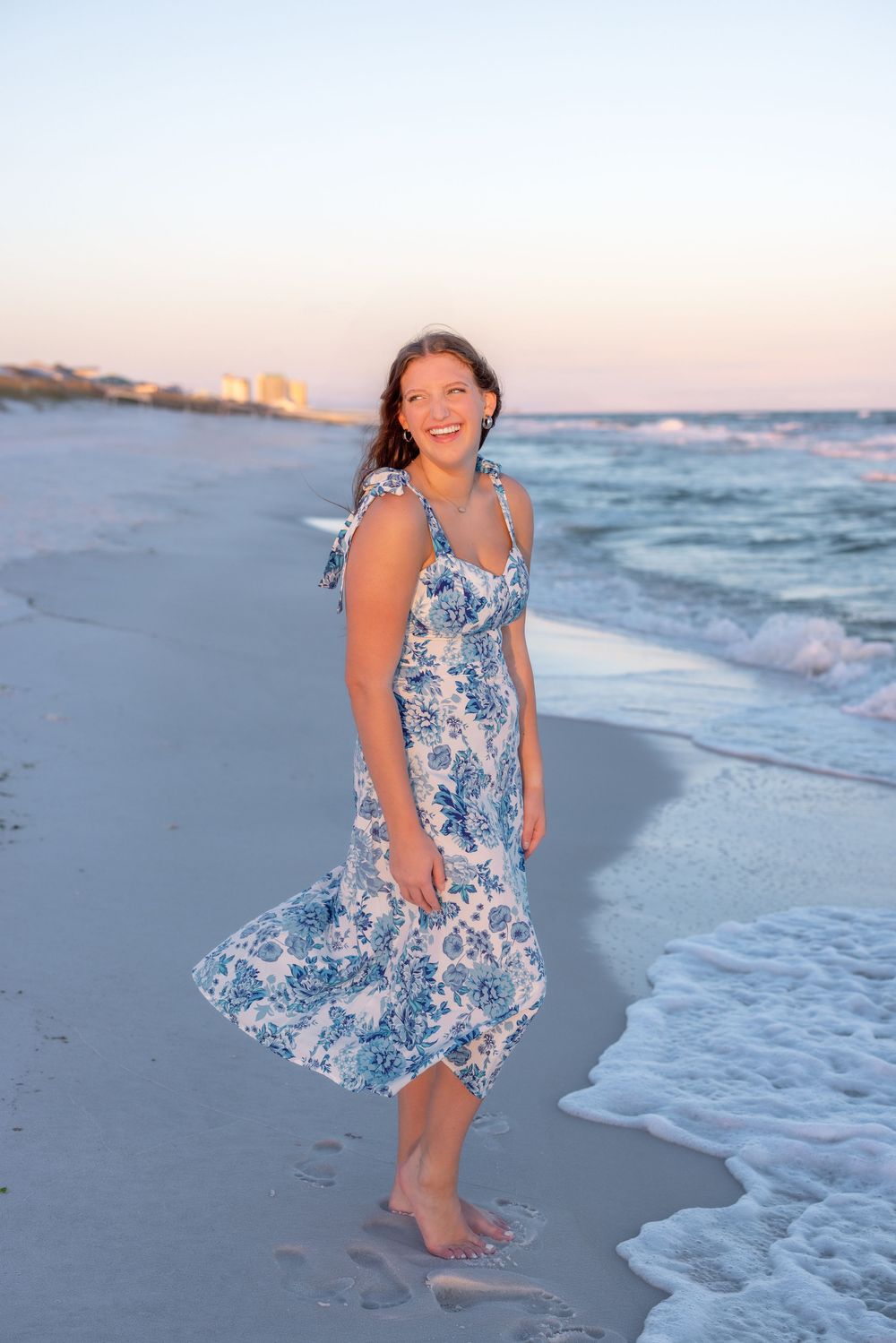 A person in a blue and white floral sundress plays joyfully at the beach during sunset.