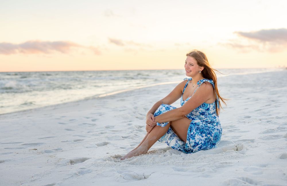 A person in a blue floral dress lounges on white sand beach at sunset creating a dreamy coastal portrait.