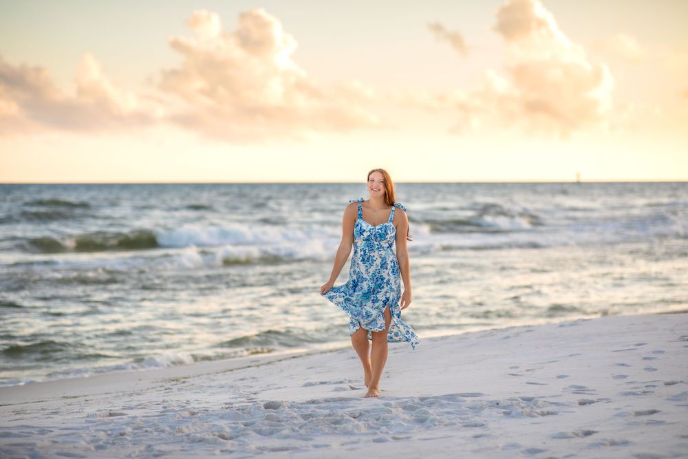 A person in a blue floral dress walks barefoot along a beautiful beach at sunset with waves crashing behind them.