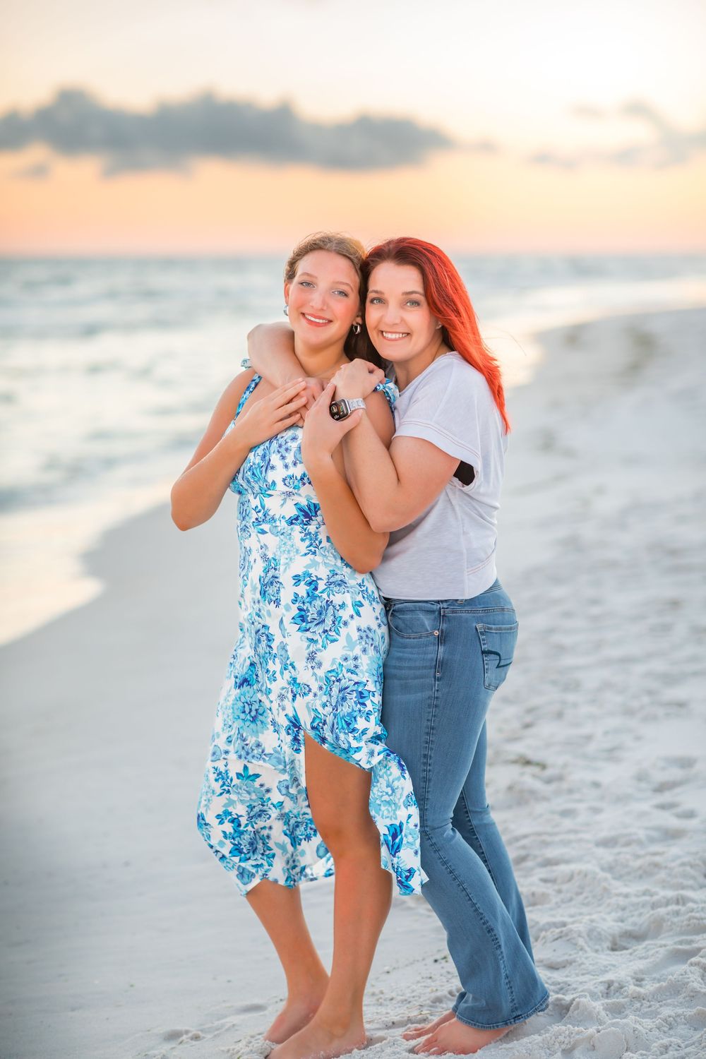 Two people embrace while laughing and playing together on a sandy beach at sunset with ocean waves in background.