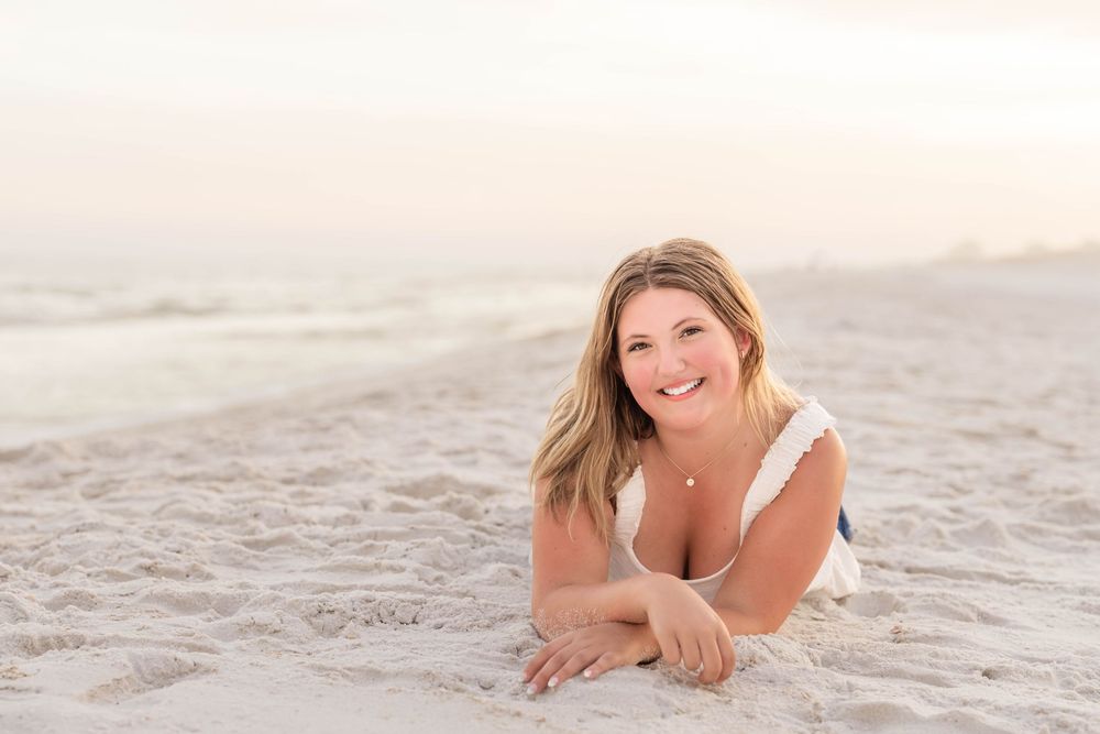 Person laying on white sand beach during sunset with soft pink sky in background.