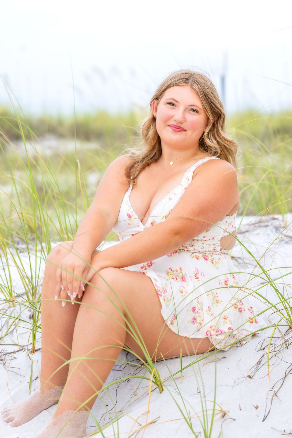 A person in a floral sundress sits smiling on a beach with sand dunes and sea grass in the background.