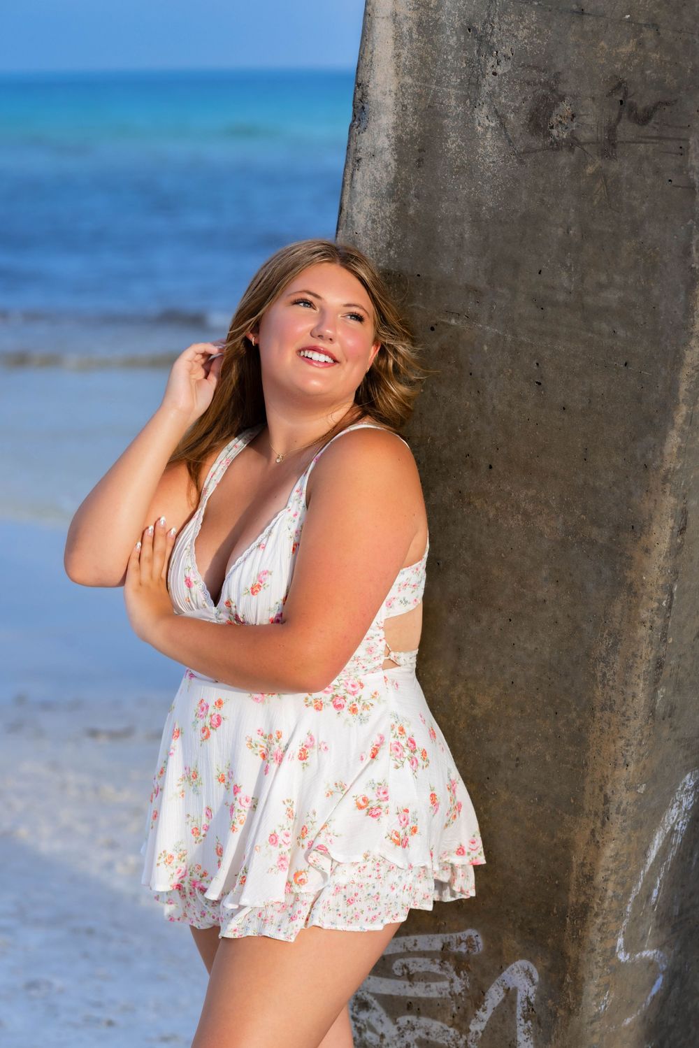 A person in a white and coral patterned sundress poses next to a palm tree at a tropical beach with blue ocean water.