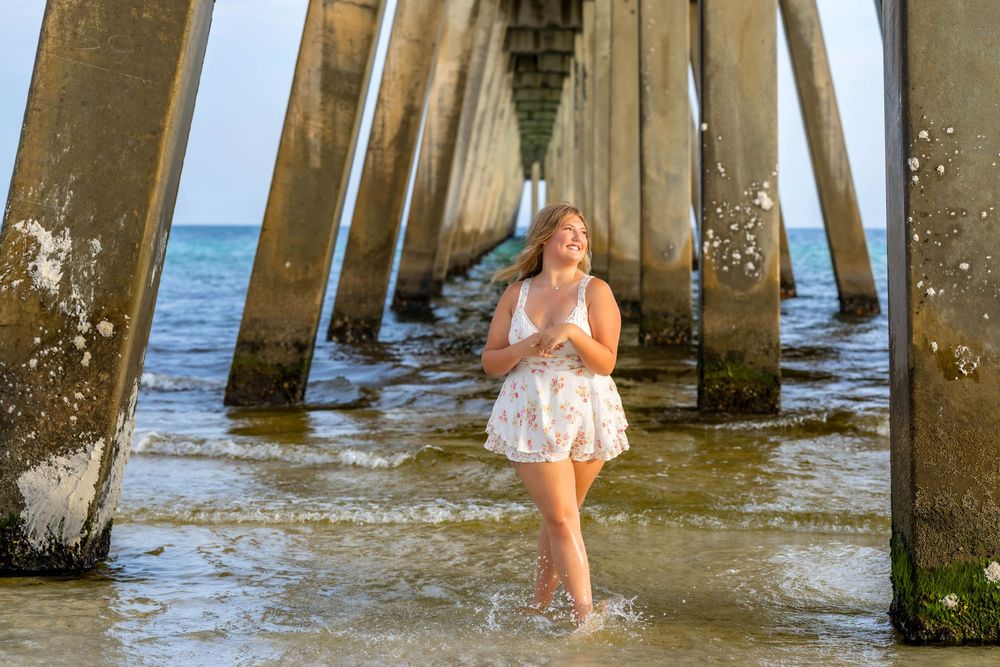 A person in a white sundress splashes in the ocean water beneath a wooden pier on a sunny day.