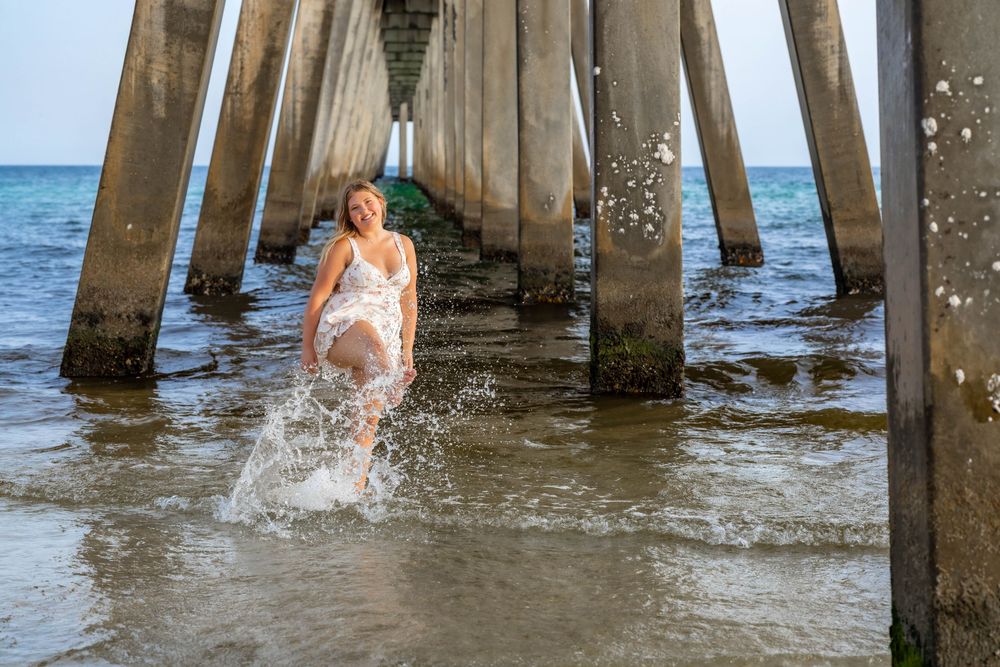Someone splashes through shallow water beneath a wooden pier on a sunny day at the beach.