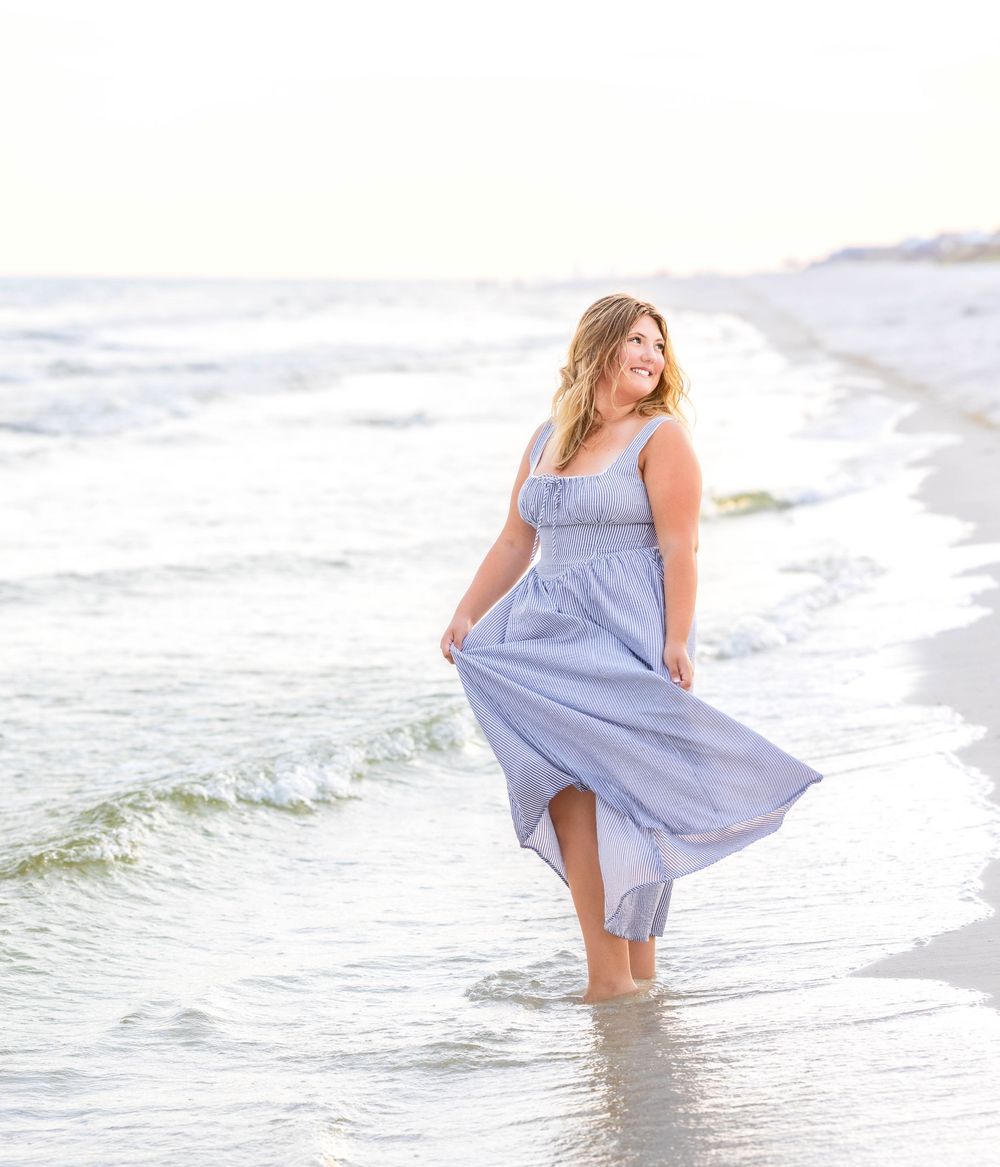 Woman in a flowing light blue dress stands at the edge of ocean waves on a sunny beach.