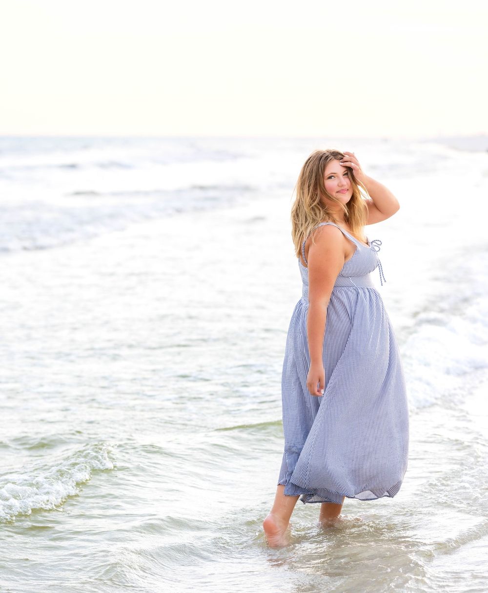 Woman in blue sundress playing and splashing in ocean waves at beach during golden hour sunset.