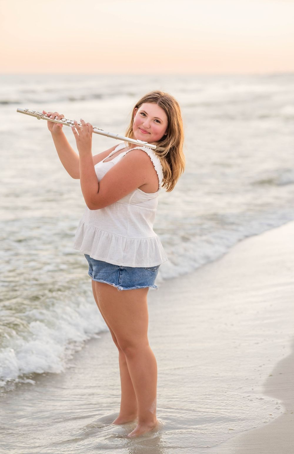 Musician poses with flute on sandy beach at sunset while standing in gentle ocean waves.