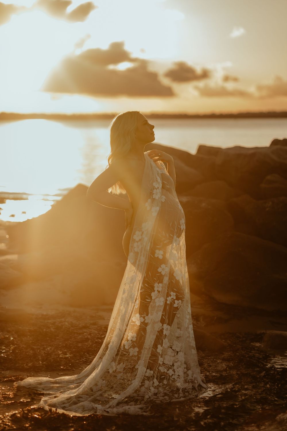 A silhouetted figure in a flowing lace dress stands on coastal cliffs at golden sunset, creating a dreamy ethereal scene.