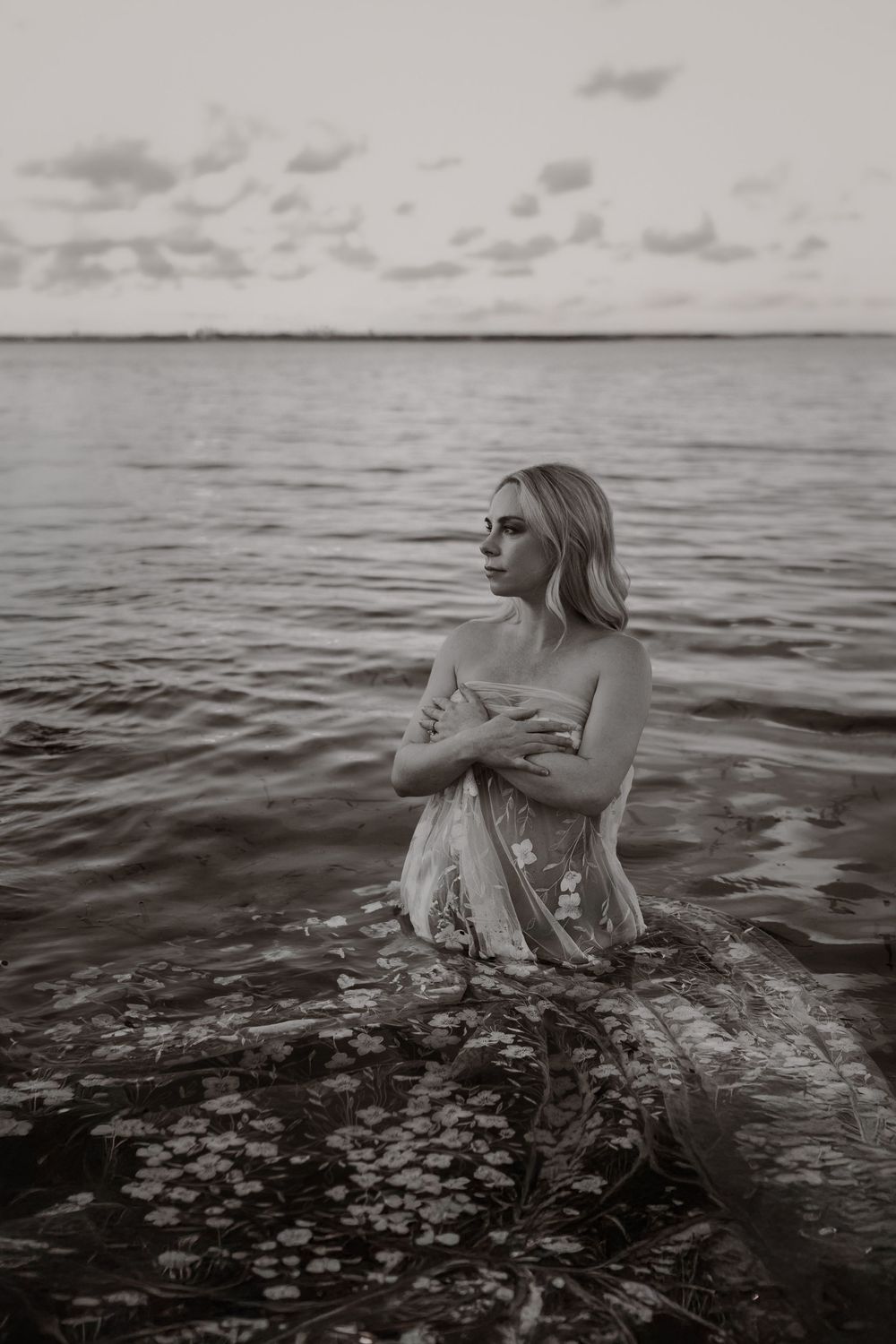 A woman in a light dress stands waist-deep in dark waters at dusk, creating a moody black and white composition.