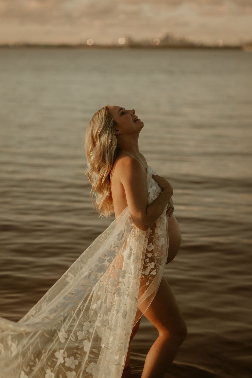 Figure in flowing white lace dress wades through serene water during golden hour, creating a dreamy atmosphere.
