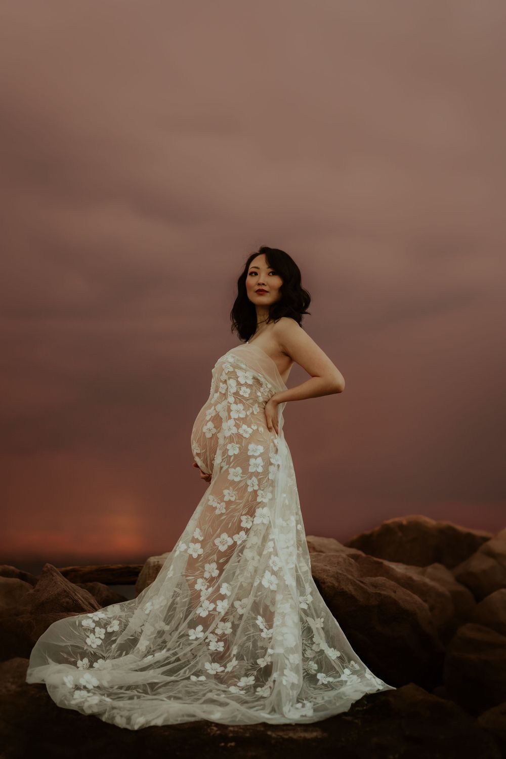 A maternity photoshoot at sunset features a woman in a flowing white lace dress standing on rocks against a dusky sky.