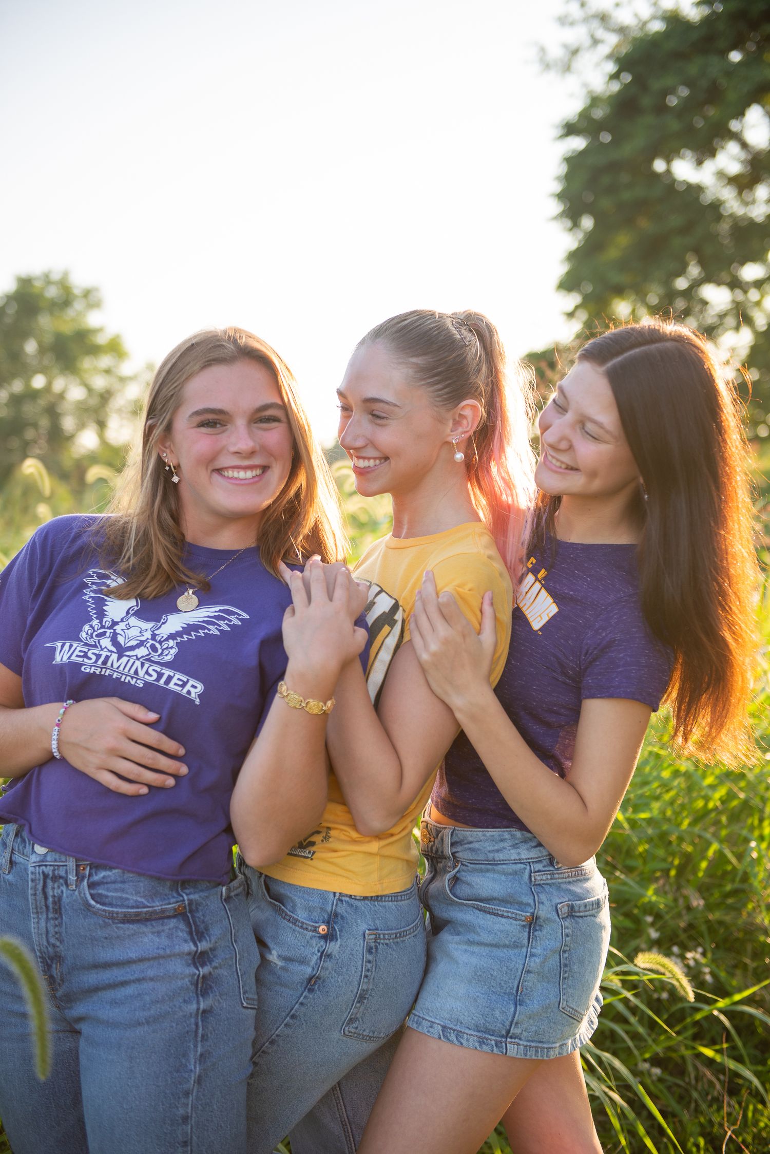 Three friends in purple and yellow shirts laughing together outdoors at sunset with golden light in the background.
