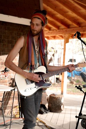 Musician wearing a colorful headband performs with an electric guitar on an outdoor wooden stage.
