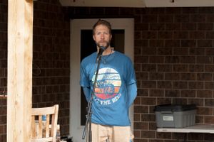 A performer in a blue t-shirt speaks into a microphone on a covered porch.