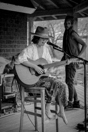 A musician performs on an acoustic guitar while sitting on a wooden stool under a covered porch.