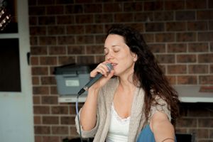 Singer performs with a microphone against a brick wall backdrop in natural lighting.