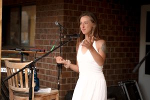 A performer in a white dress speaks into a microphone against a brick wall backdrop.
