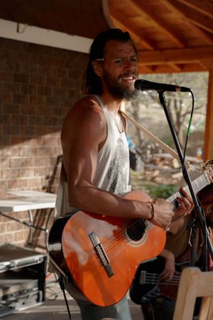Musician performs with an orange acoustic guitar under a covered porch with brick walls and wooden beams.