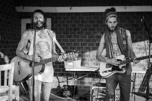 Black and white photo of two musicians performing with guitars against a brick wall backdrop.