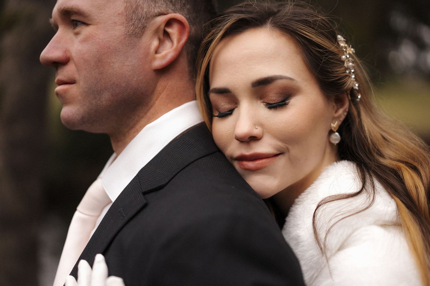 Close up of a bride and groom standing together at an outdoor wedding, with bride resting her eyes behind groom.