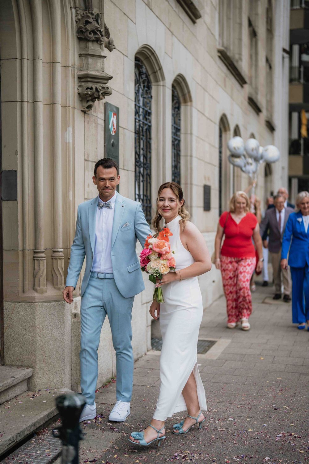 Couple in light blue suit and white dress pose with flowers outside historic building.