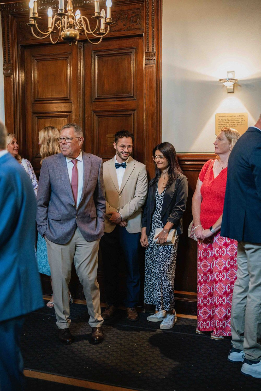 Wedding guests gather in formal attire inside wood-paneled room.