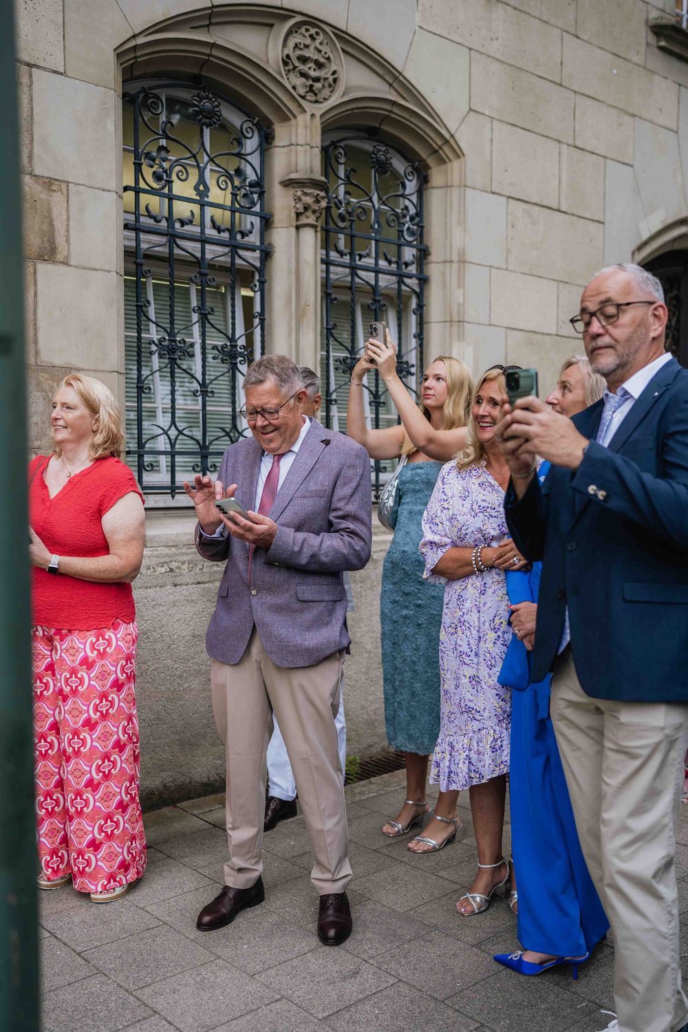 Wedding guests in formal attire applaud while standing along a stone building wall.