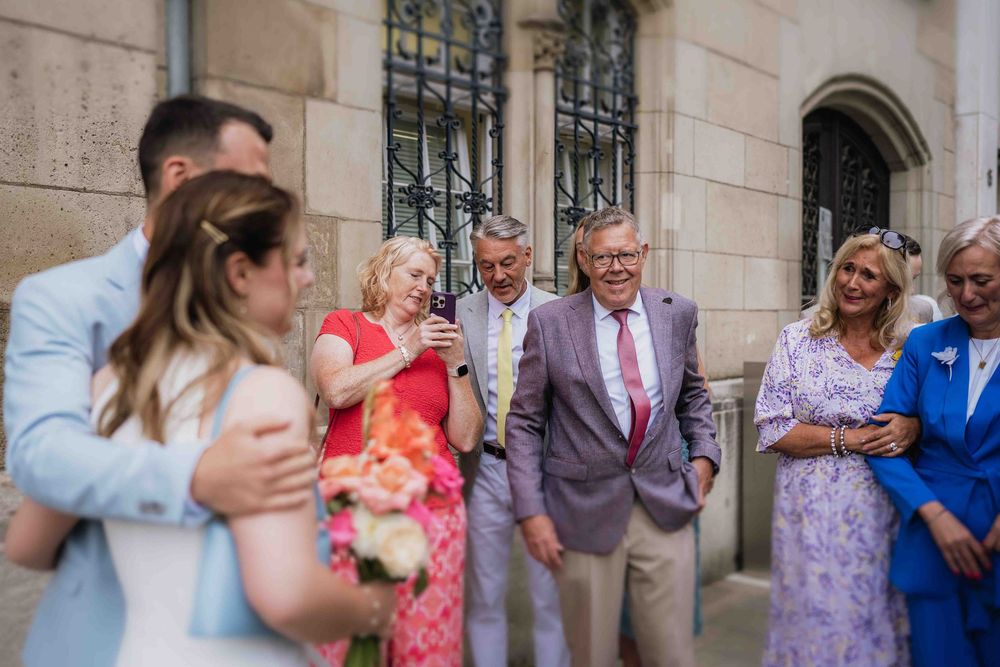 Wedding guests in colorful attire gather along a stone building to celebrate.