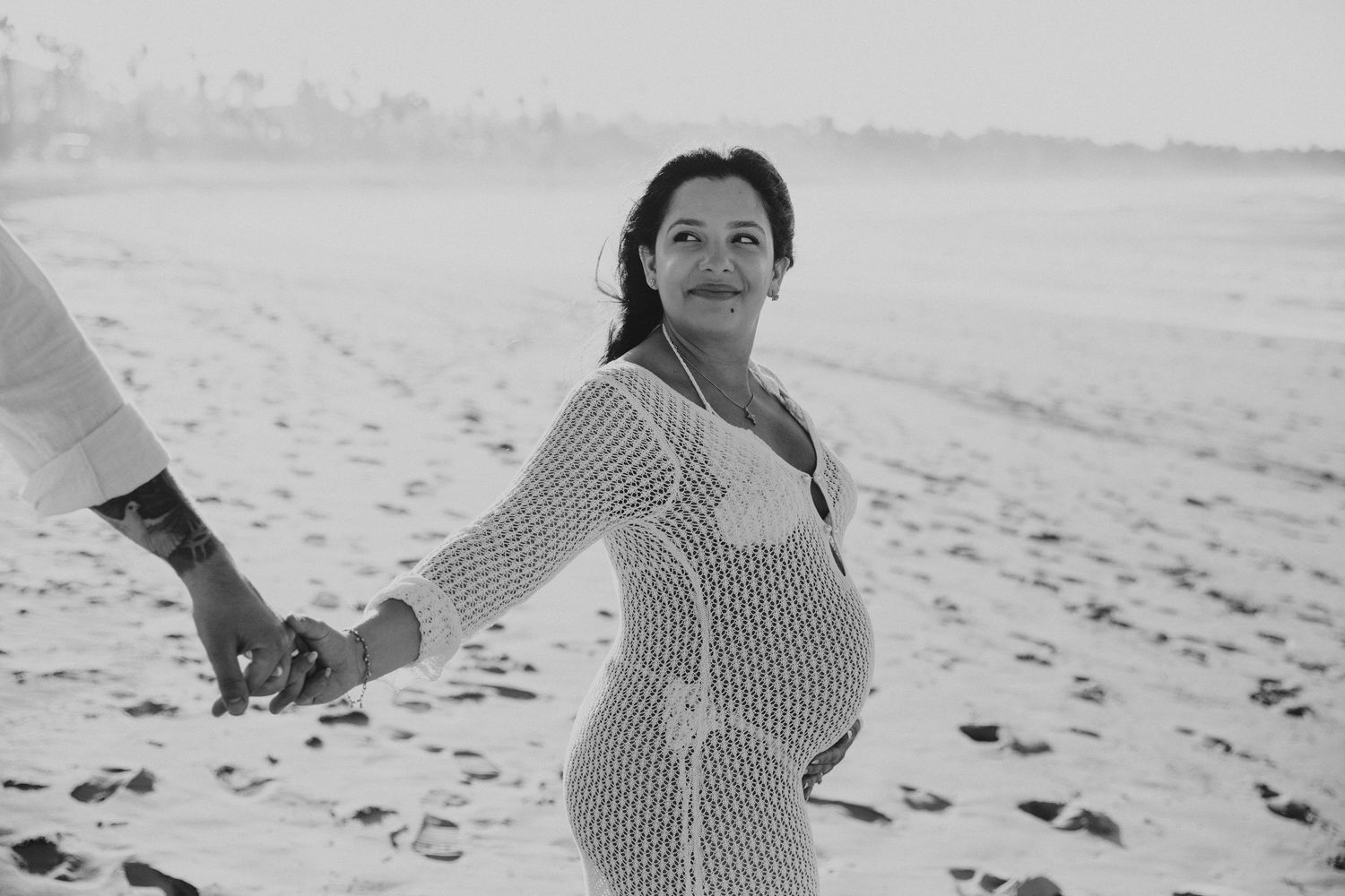 A black and white photo of a woman in a crochet dress walking on a beach.