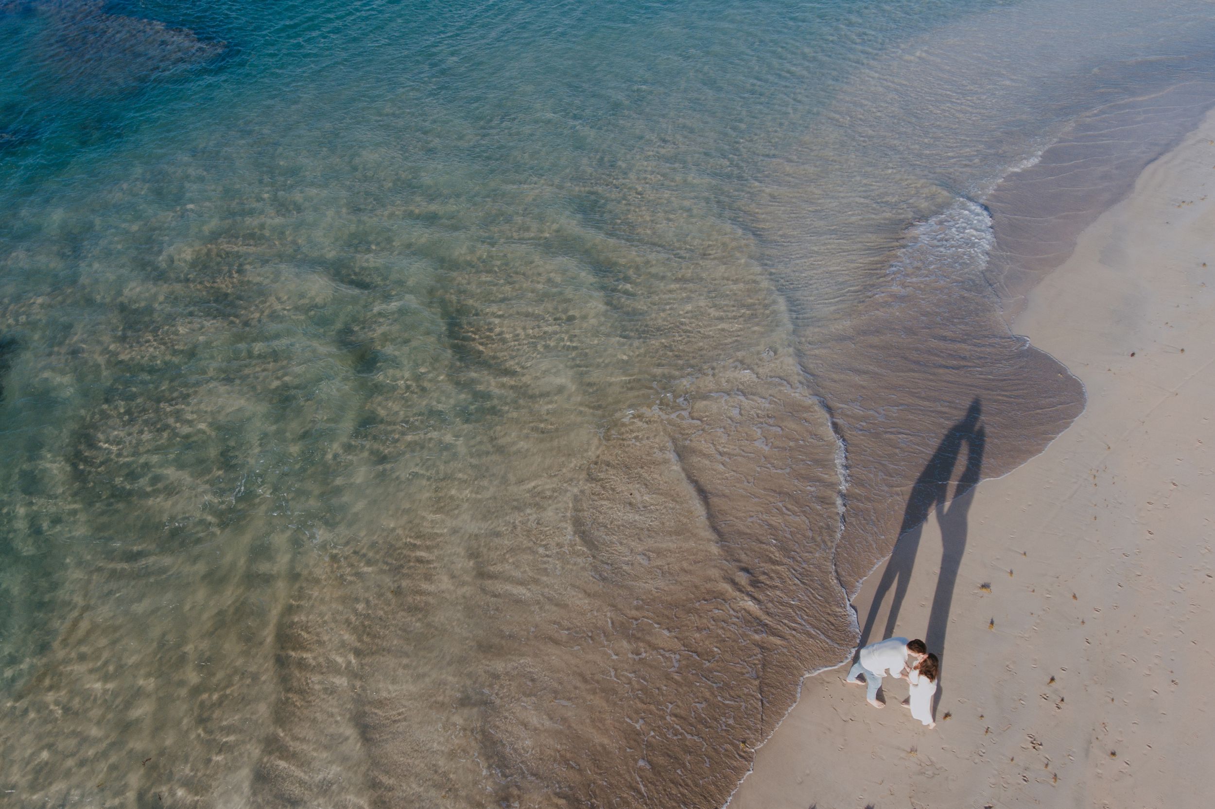 Aerial view of a beach scene with crystal clear waters and long shadows cast on the sand.