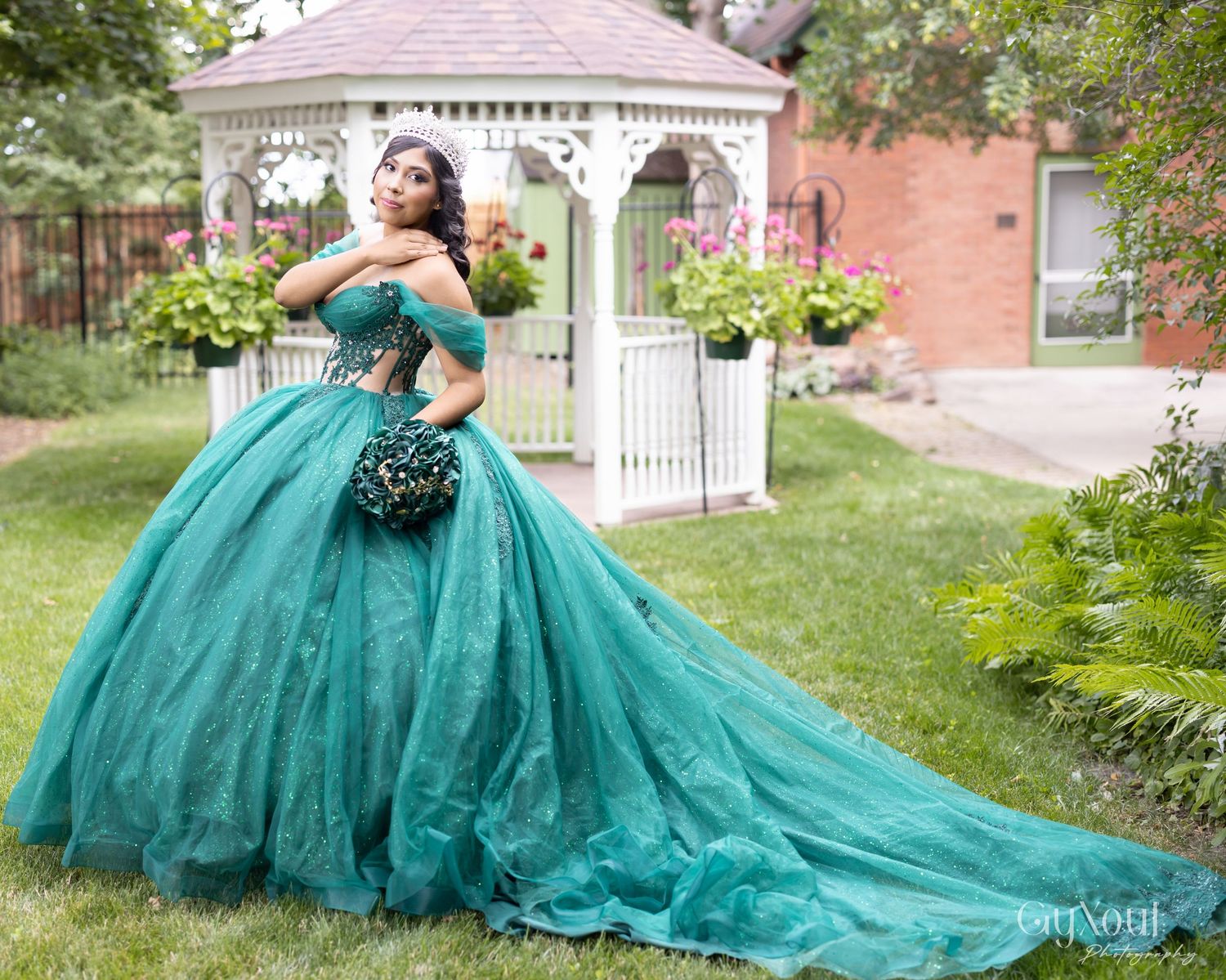 A stunning teal quinceañera ball gown with a flowing train photographed in front of a white garden gazebo.