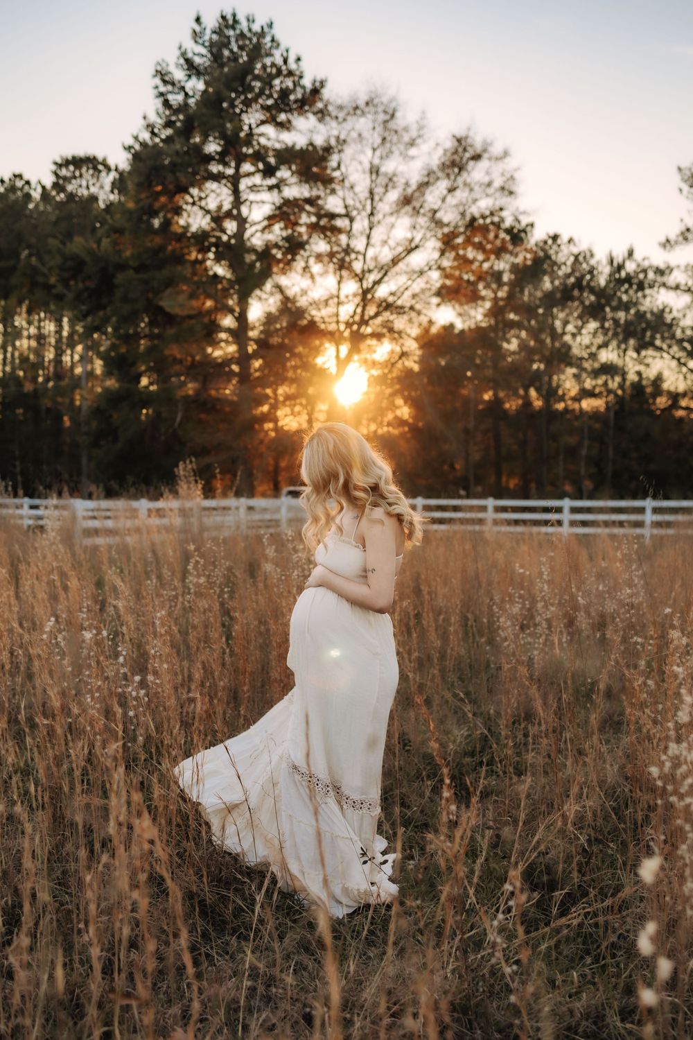 A couple shares a tender moment beside white fencing on a grassy hill at sunset in a pastoral countryside setting.