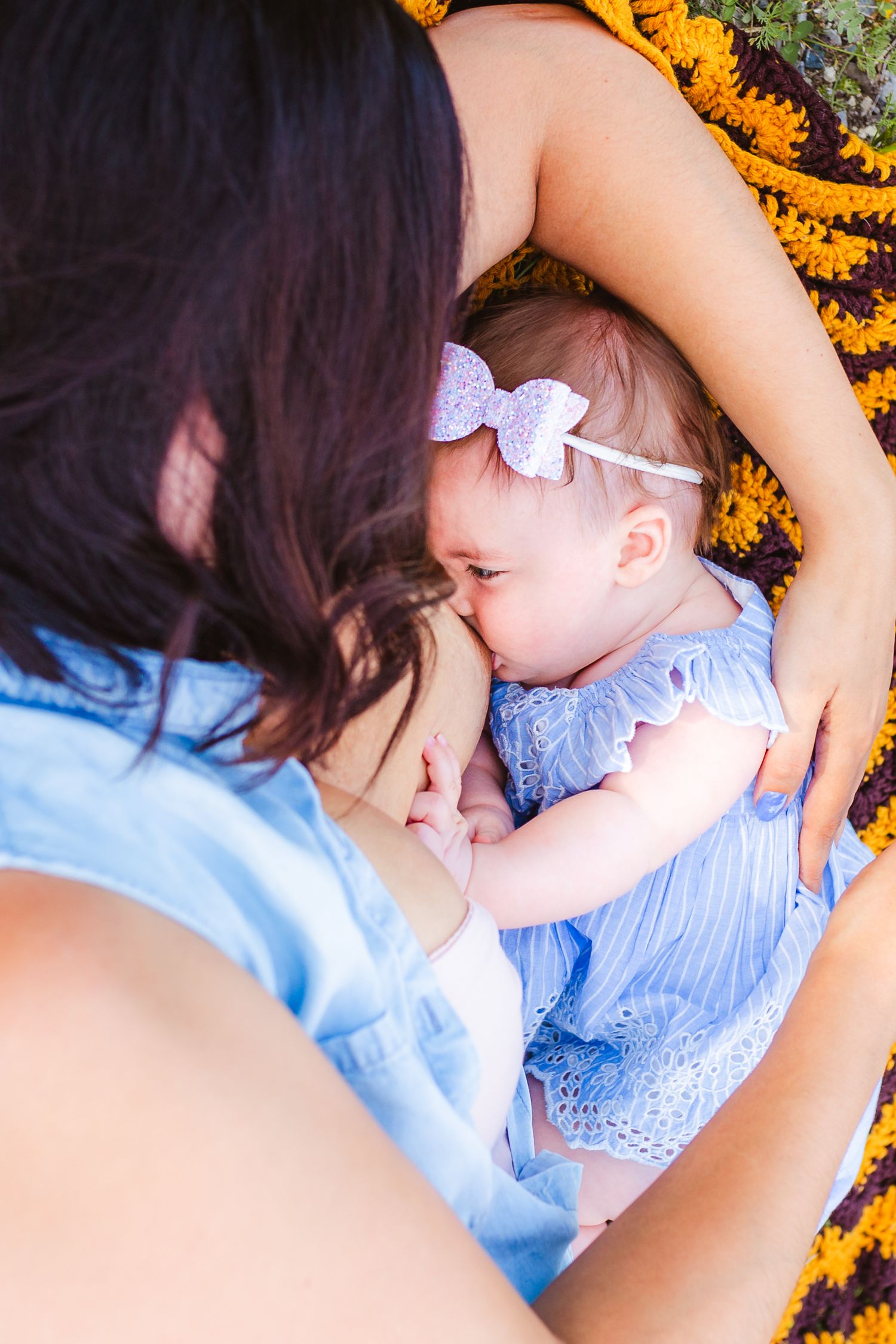 Mother wearing blue dress breastfeeding baby while lying on orange and brown blanket.