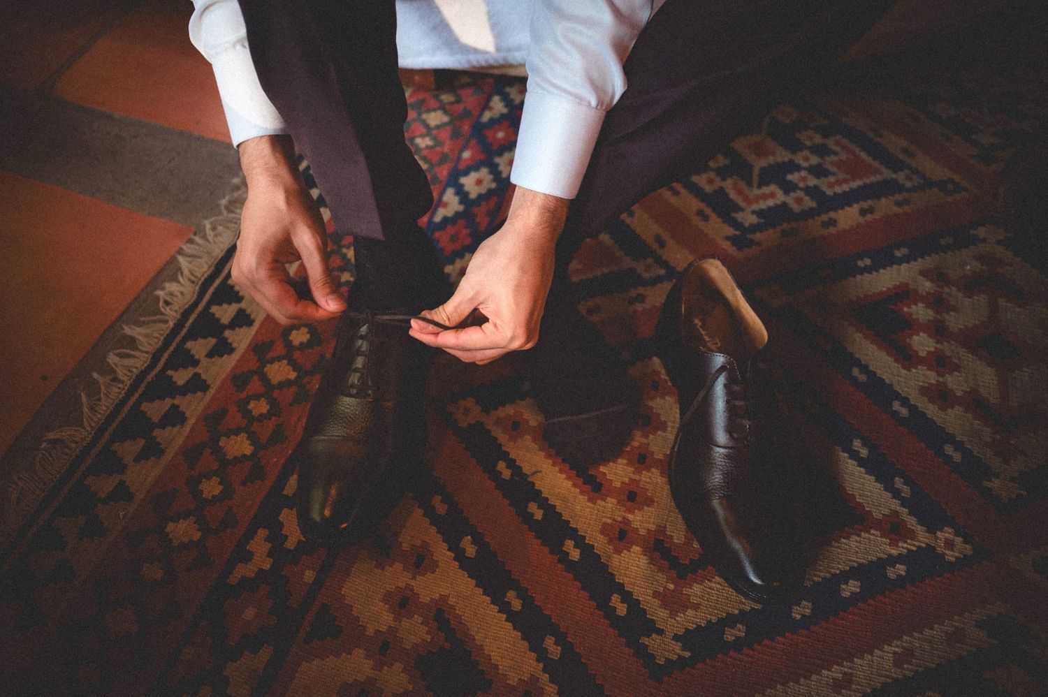 Someone adjusts formal footwear on an ornate vintage carpet with patterned designs in warm colors.