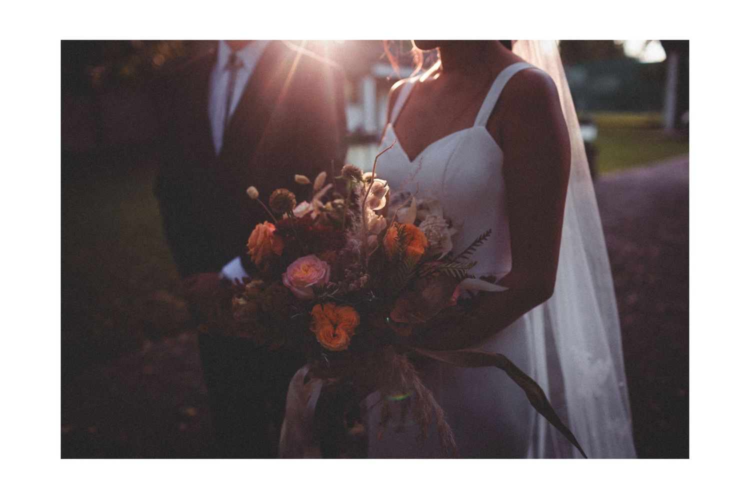 A sunlit wedding photograph showing a white bridal gown and romantic burgundy floral bouquet at golden hour.