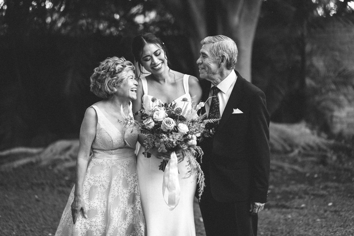 A vintage black and white wedding photograph showing a joyful moment between three people in formal attire outdoors.