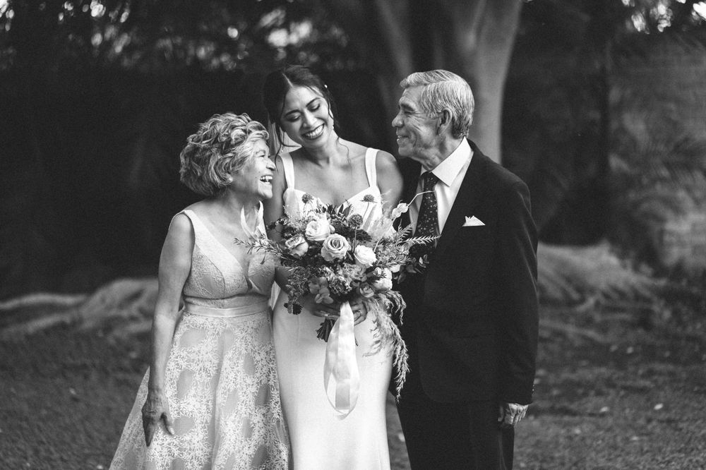 A vintage black and white wedding photograph showing a joyful moment between three people in formal attire outdoors.