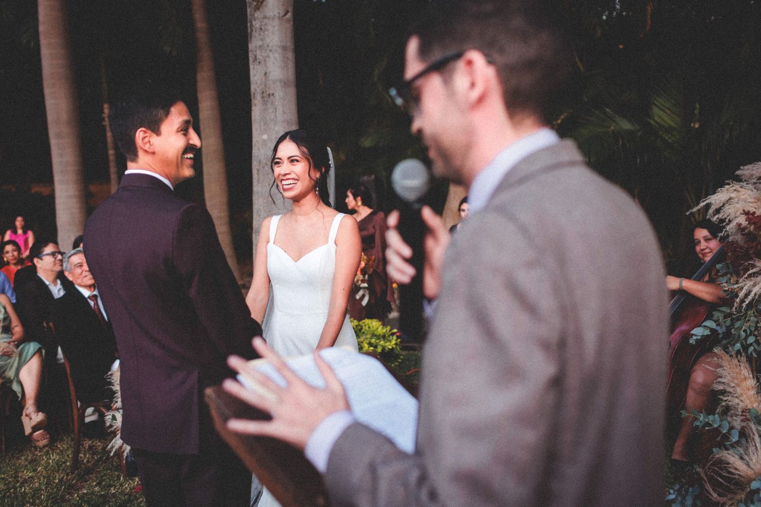 An outdoor wedding ceremony at dusk shows a bride and groom exchanging vows with the officiant speaking into a microphone.