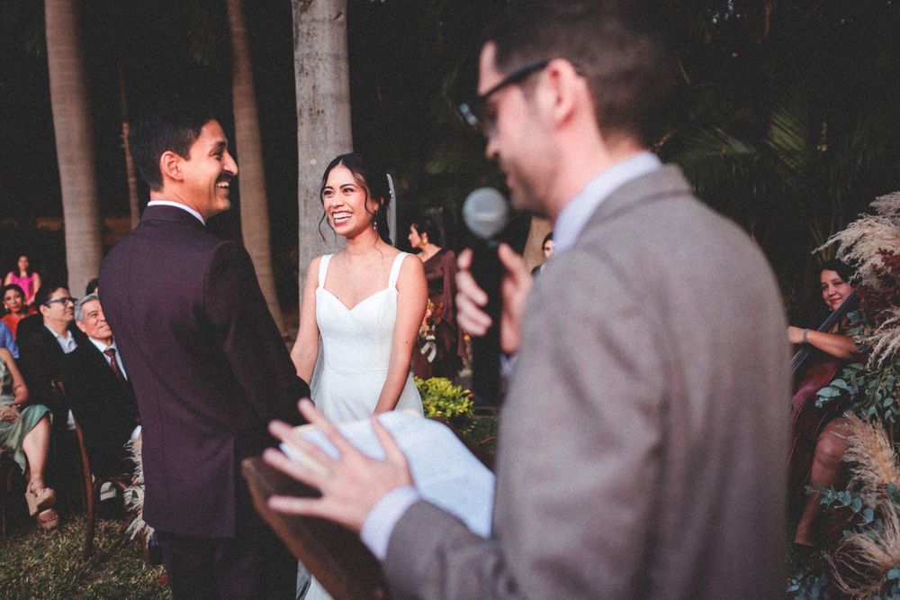 An outdoor wedding ceremony at dusk shows a bride and groom exchanging vows with the officiant speaking into a microphone.