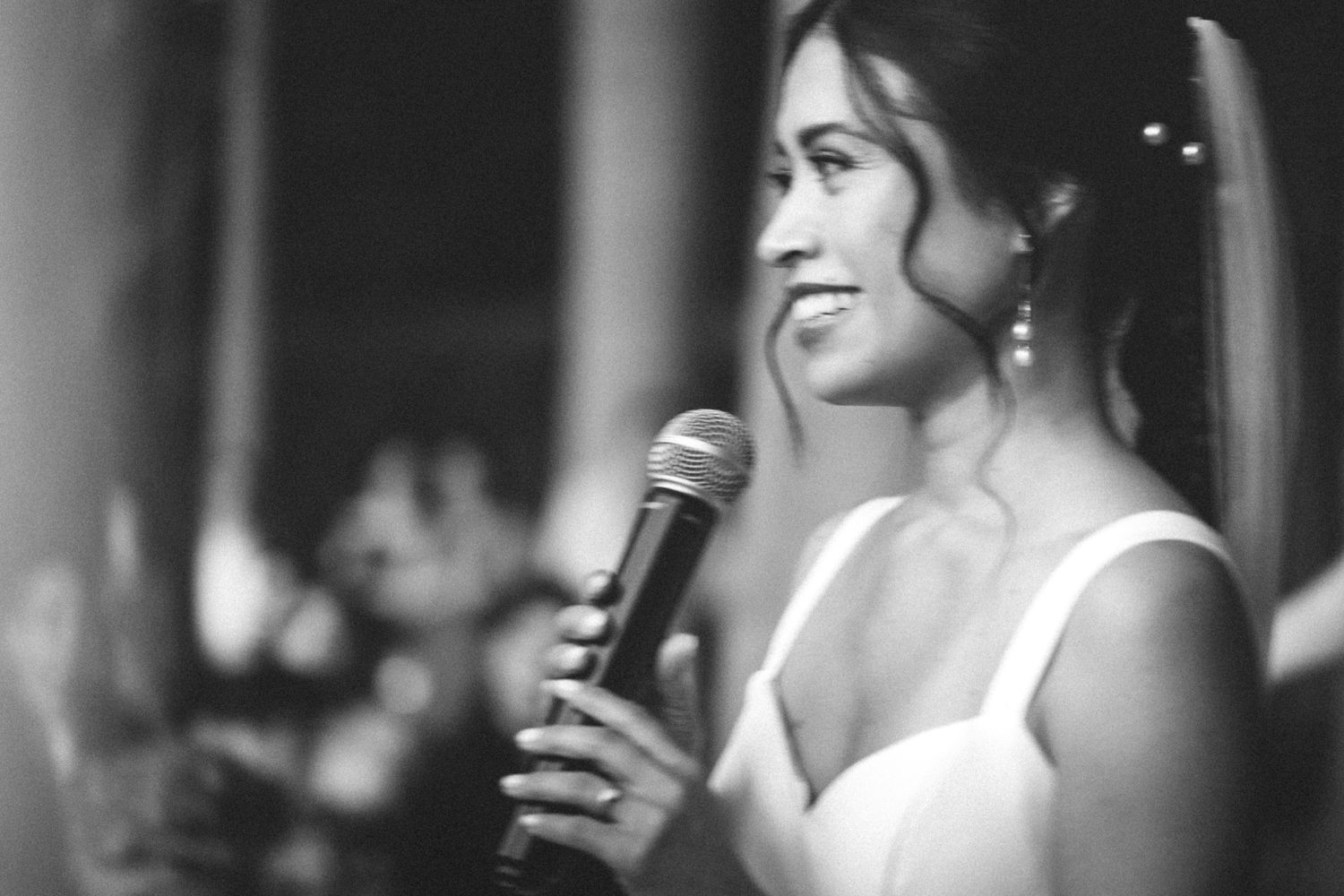 Black and white photo of someone speaking with a microphone in an event setting with a sleeveless white dress.